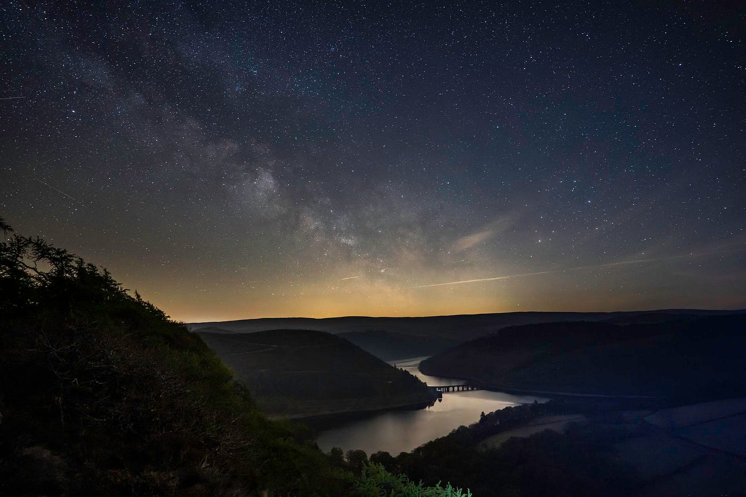 A breathtaking night photograph of the dense Milky Way galaxy stretching across a brilliant, star-filled dark sky over the Elan Valley. The dark silhouettes of rolling hills frame a calm, winding reservoir reflecting the starlight, with the stone arches of the Pont ar Elan bridge visible in the distance.