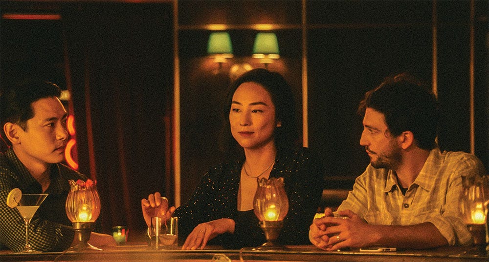 Três pessoas sentadas em um bar à meia-luz, um homem e uma mulher de ascendência coreanas e um homem branco. eles estão contemplativos e reflexivos