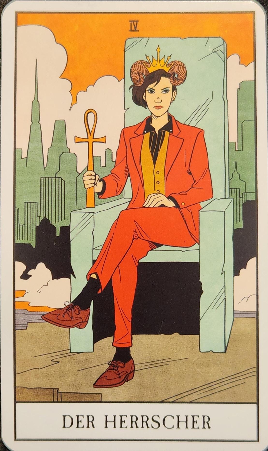 A tarot card labelled IV Der Herrscher. A non-binary presenting white person is sitting outdoors on a large throne, that appears to be made of either glass or stone. They are wearing a red suit, with a beige vest and a black shirt. On their head is a golden crown, nestled between two ram horns. They are sitting cross-legged and holding a large golden ankh. Behind them is the skyline of a large city, covered in mist and clouds.