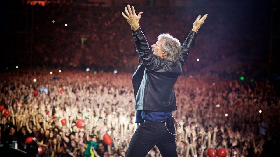 Jon Bon Jovi, wearing a black leather jacket and black jeans, stands with arms aloft facing a huge crowd. He's singing Livin' on a Prayer Jon Bon Jovi, wearing a black leather jacket and black jeans, stands with arms aloft facing a huge crowd. He's singing Livin' on a Prayer