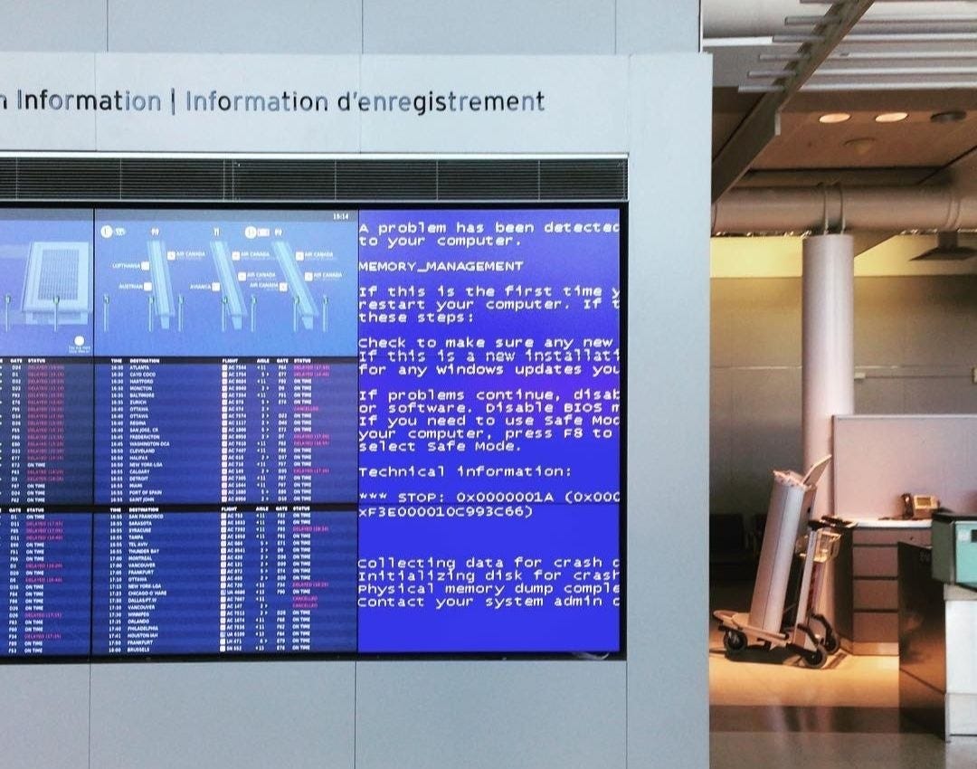 A Canadian airport departures board showing a Windows blue screen of death (MEMORY_MANAGEMENT error) on one of its display panels, next to the flight status listings. The BSOD shows a STOP error with a physical memory dump in progress.