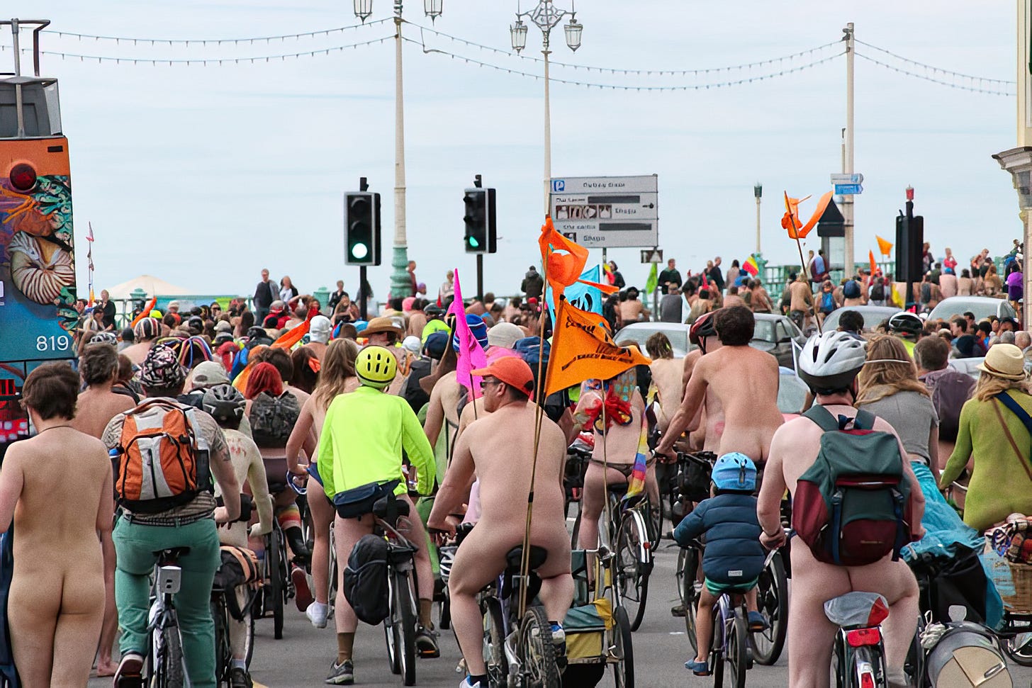 A large crowd of cyclists, many nude, rides along King's Road in Brighton under an overcast sky. Riders wear helmets in bright colors and carry orange flags.