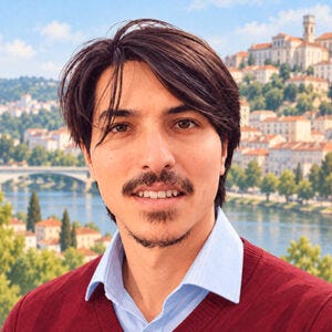 Fotografia de um homem jovem, de cabelos castanhos escuros, lisos e médios, bigode e barba rala no queixo. Ele sorri para a câmera e veste uma camisa azul-clara sob um suéter vinho com decote em V. Fotografia de um homem jovem, de cabelos castanhos escuros, lisos e médios, bigode e barba rala no queixo. Ele sorri para a câmera e veste uma camisa azul-clara sob um suéter vinho com decote em V.