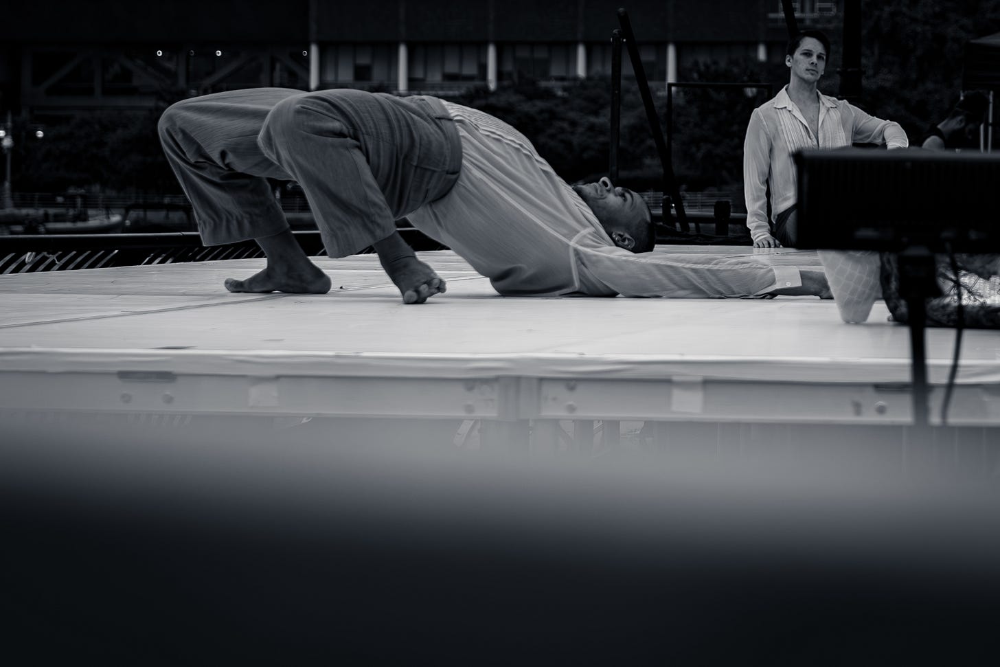 A dancer lies arched on their back on stage, while another performer looks on from the side of the platform. A dancer lies arched on their back on stage, while another performer looks on from the side of the platform.