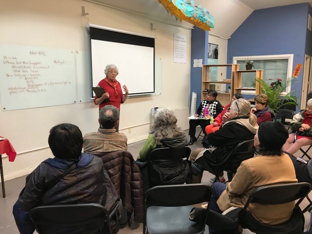 An older woman in red speaking to a seated group of people.