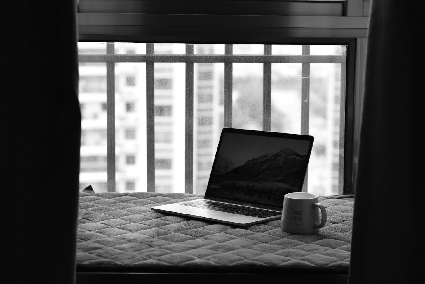 A black-and-white photo of an open laptop and a mug on a cushioned window seat in front of a bright window. A black-and-white photo of an open laptop and a mug on a cushioned window seat in front of a bright window.