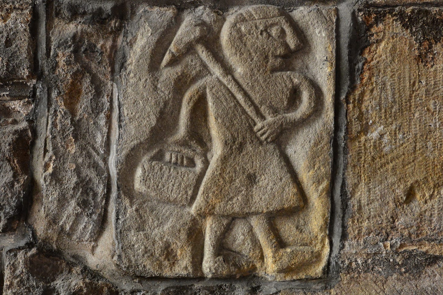 Stone carving of a person in a tunic carrying a tool over their shoulder and holding a basket, set into a rough stone wall. Stone carving of a person in a tunic carrying a tool over their shoulder and holding a basket, set into a rough stone wall.