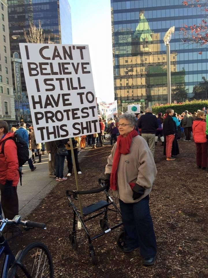 Mulher idosa ao lado de um andador onde está amarada uma haste de uma placa grande com o escrito: "I can’t believe I still have to protest this shit" (tradução livre: Não consigo acreditar que ainda tenho que protestar por essa merda de assunto)