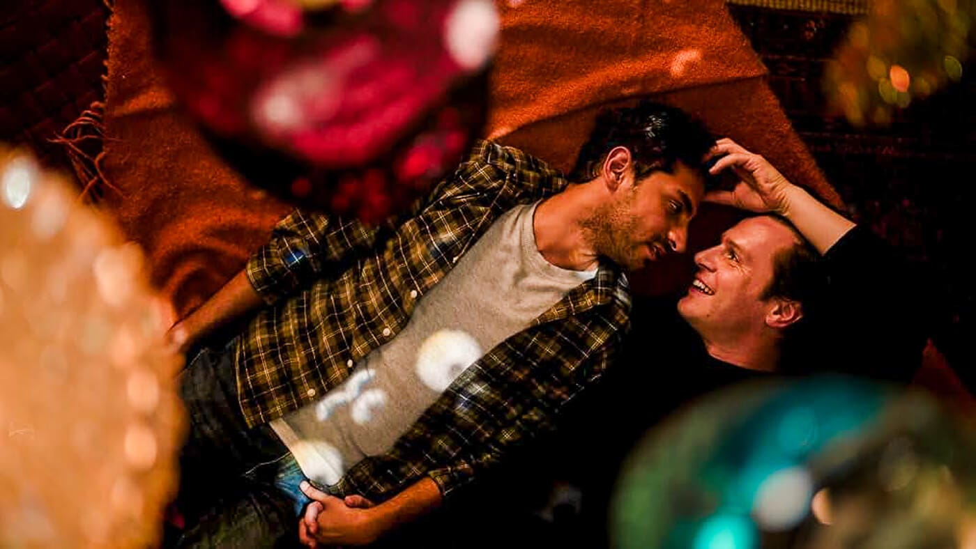 Naveen and Jay lie face to face on a rust-colored rug, surrounded by warm overhead ornaments and soft shadows.