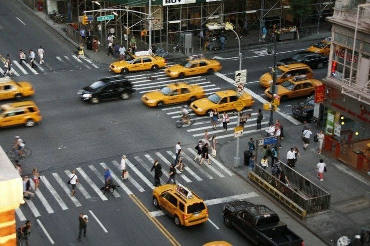 This may contain: a busy city street filled with lots of yellow cabs and people crossing the street