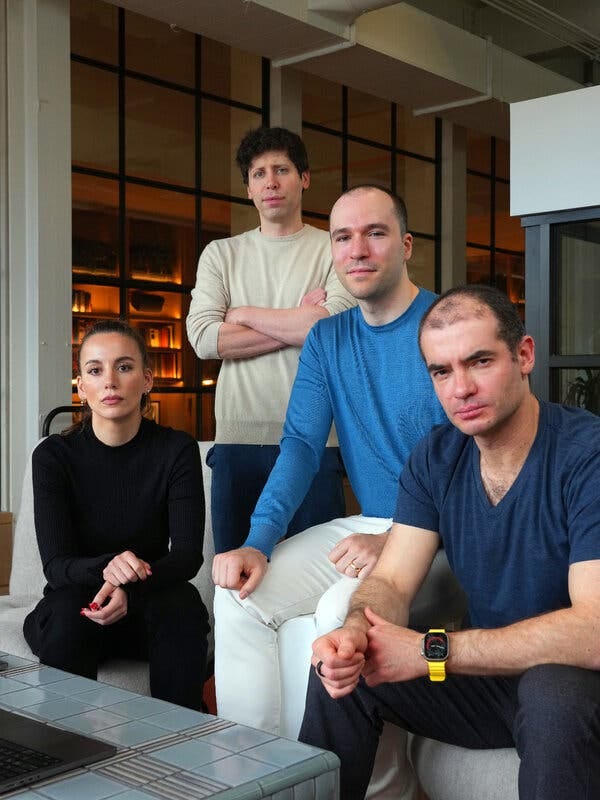 Ms. Murati and two men sit next to one another on upholstered white chairs in an office. Mr. Altman stands behind them with his arms crossed. All are looking at the camera. 