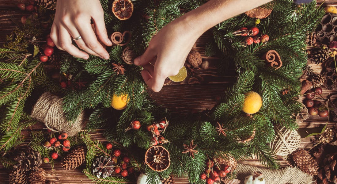 A pair of hands decorating a pine leaf wreath, with oranges, berries, and lemons around it