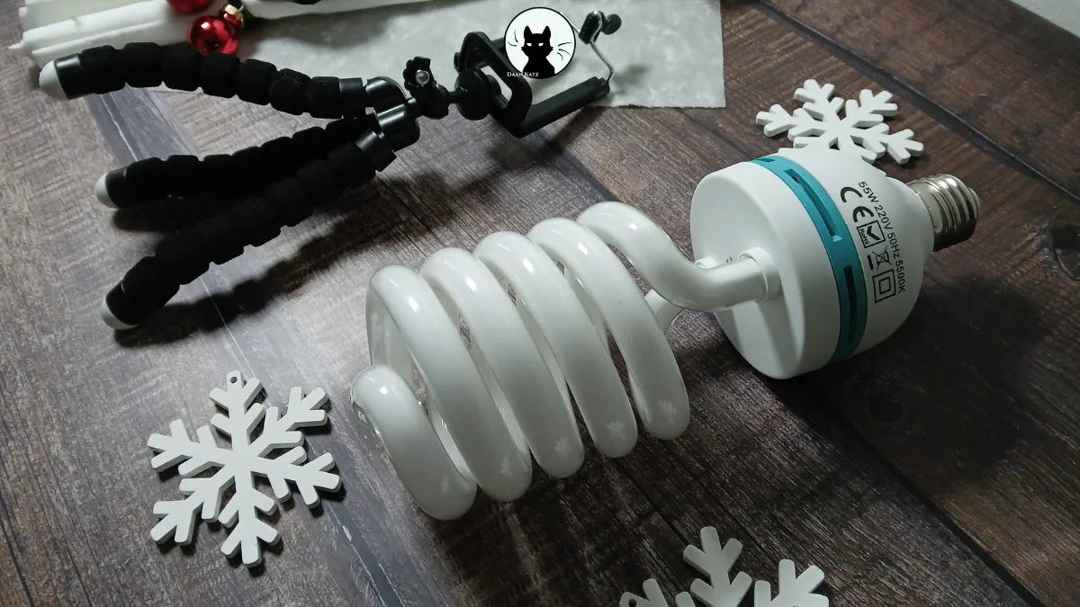 A spiral CFL studio light bulb lies on a wooden surface beside white snowflake decorations and a black flexible tripod and decorative red baubles, in a compact studio setup.