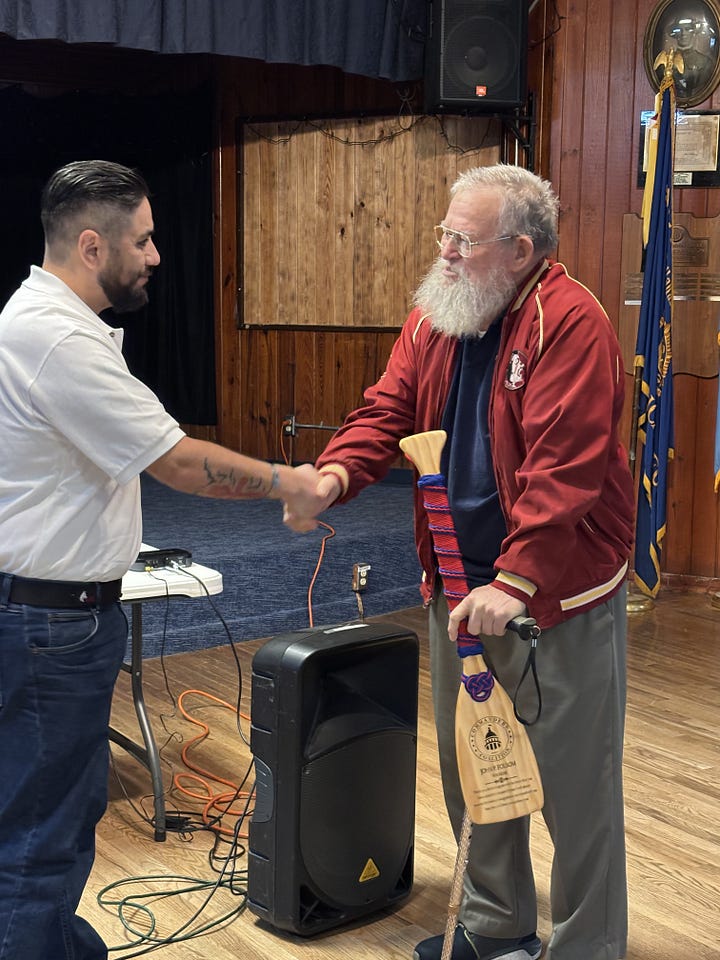 Two men shake hands in a wood‑paneled hall near a small stage. The man on the left wears a white polo and jeans; the older man on the right, with a white beard and glasses, wears a red jacket and gray slacks and leans on a decorative cane wrapped with red‑and‑blue cord. A small paddle attached to the cane bears an insignia with a dome resembling the U.S. Capitol and the words “Commanders Coaliiton.” A black Behringer PA speaker sits between them, with audio cables running to a folding table holding equipment. A blue flag with gold fringe and framed plaques hang on the wall in the background.