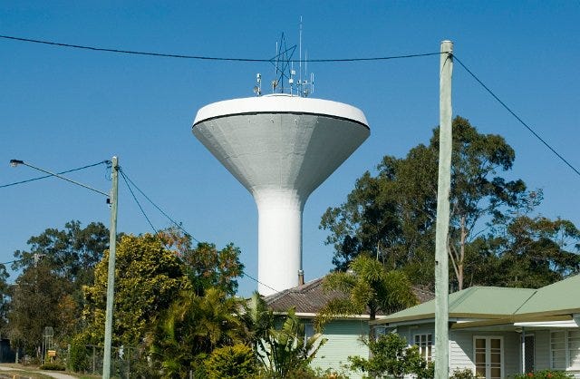 White concrete funnel-shaped water tower looming in the background above some houses and trees on a cloudless day