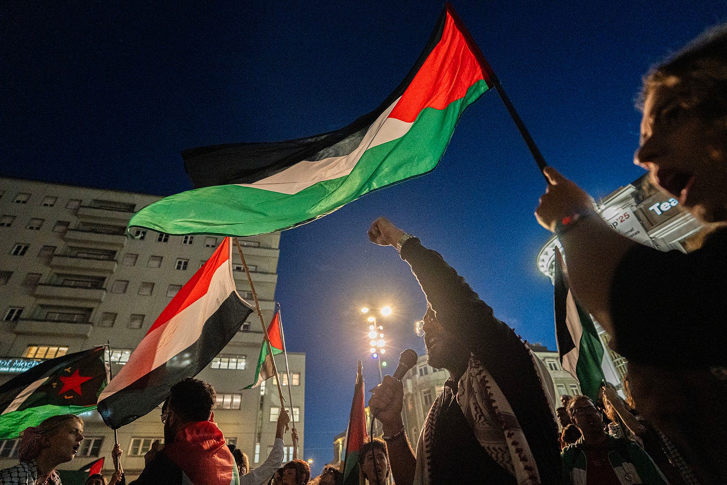 Una fotografia notturna di una manifestazione con una folla di persone, illuminata dai lampioni e dalle finestre degli edifici vicini. Sono visibili diverse grandi bandiere, principalmente la bandiera palestinese (strisce nere, bianche, verdi e rosse con un triangolo rosso), tenuta in alto e che sventola al vento. Un uomo in primo piano al centro, che indossa una kefiah, sta alzando il pugno in aria e sembra parlare in un microfono. 