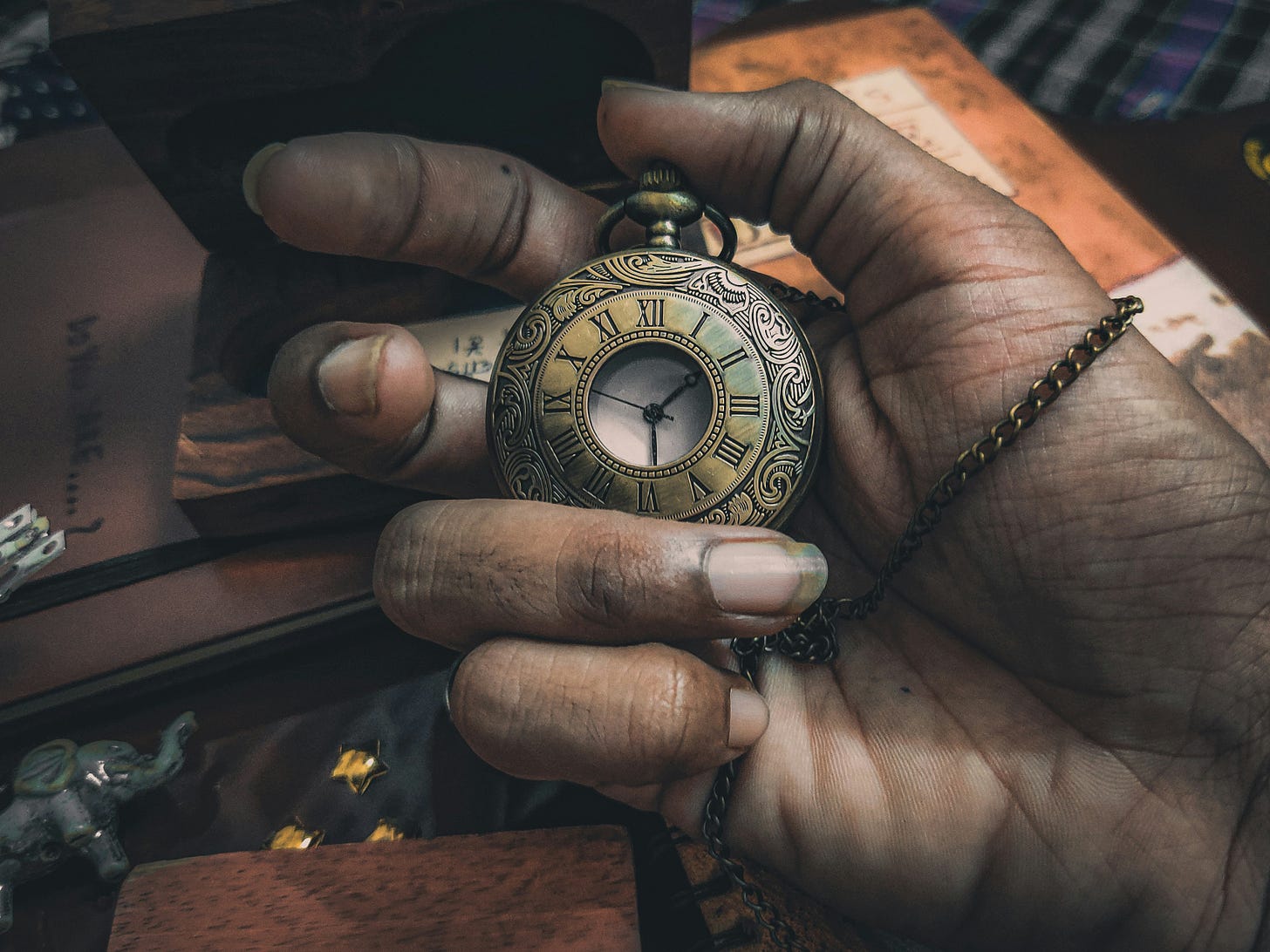 A hand holding a golden pocket watch with a desk and books in the background.