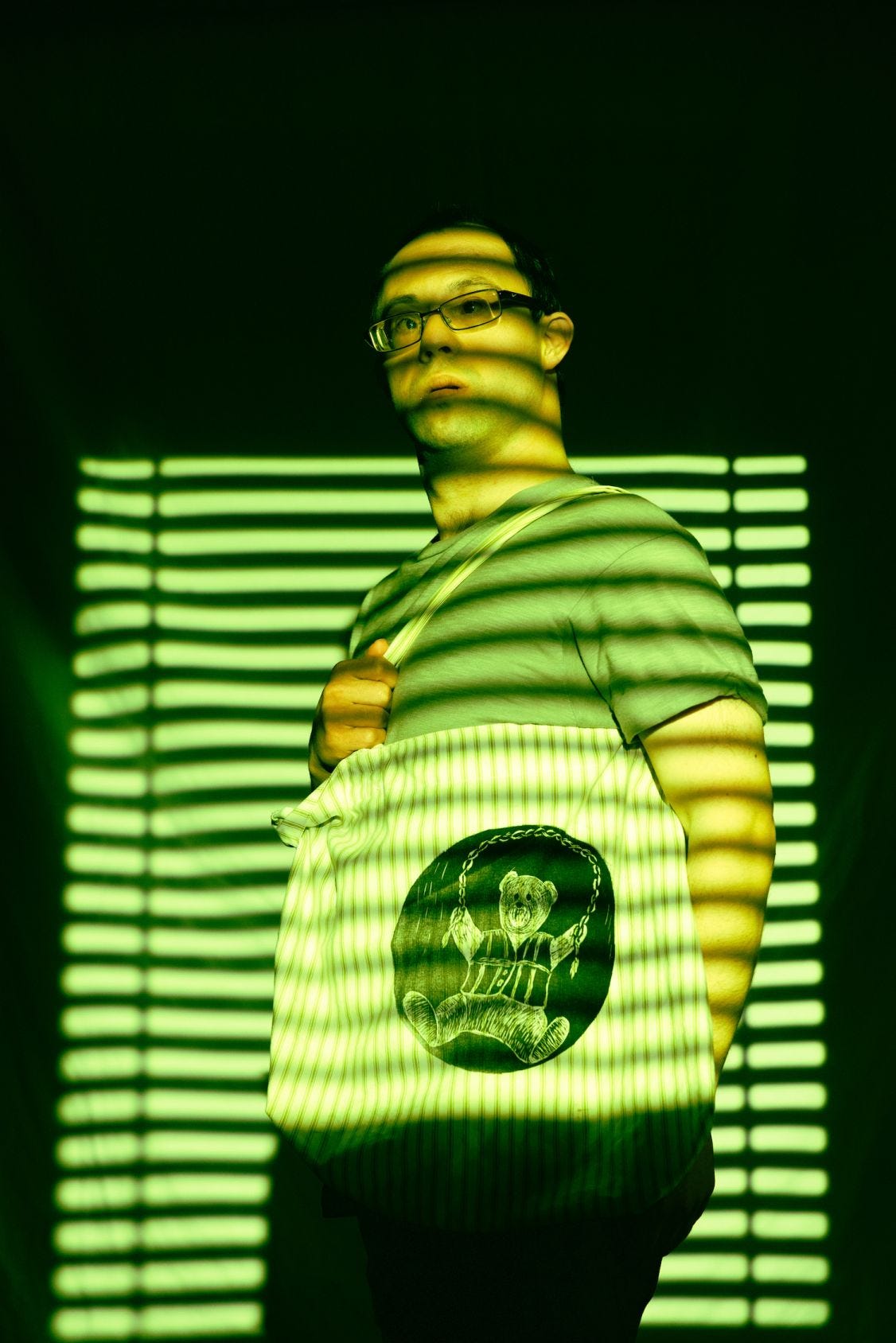 A man with Down syndrome is bathed in green light that seems to be coming through the slats of window blinds. He holds a tote bag with a drawing of a teddy bear.