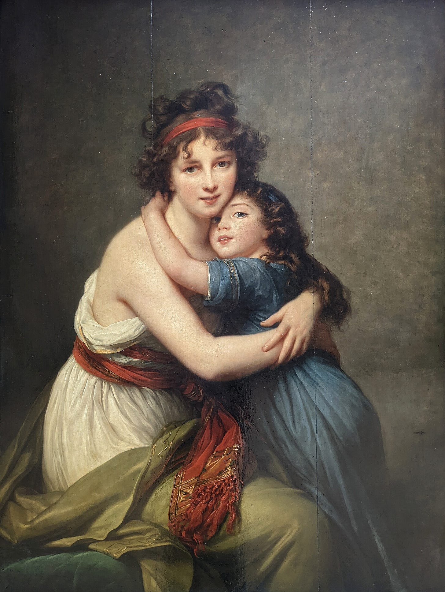 A young mother in a pale dress and headscarf holds her small daughter tightly in her arms, both turned toward the viewer, the mother smiling with her lips slightly parted. A young mother in a pale dress and headscarf holds her small daughter tightly in her arms, both turned toward the viewer, the mother smiling with her lips slightly parted.