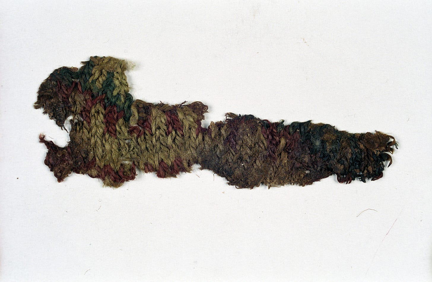 A long, irregular piece of deteriorated medieval knitting, brownish-red in colour, showing fragments of stranded colourwork. The fabric is frayed at the edges but the stitch structure is partially visible