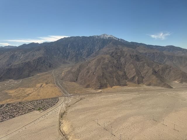 Aerial view of the arid desert landscape near Palm Springs, California, with winding roads, a residential neighborhood in the lower left, and the towering, rugged San Jacinto Mountains in the background, topped with a light dusting of snow.