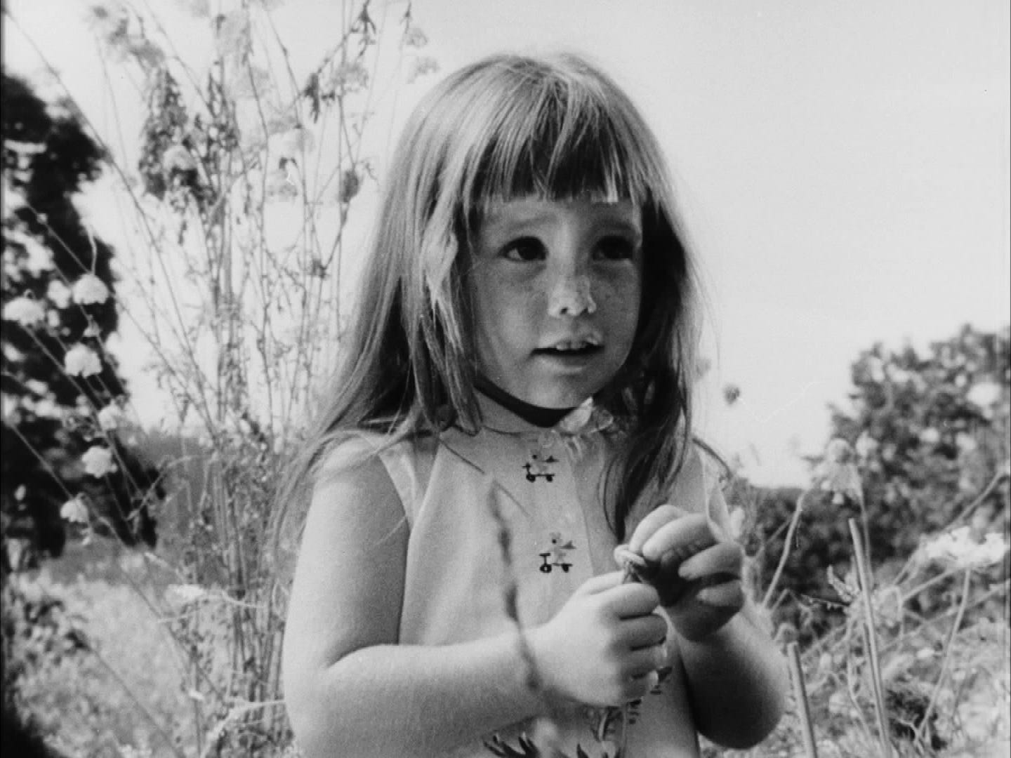 Photo of young white girl with long hair wearing a sleeveless dress and holding a flower.