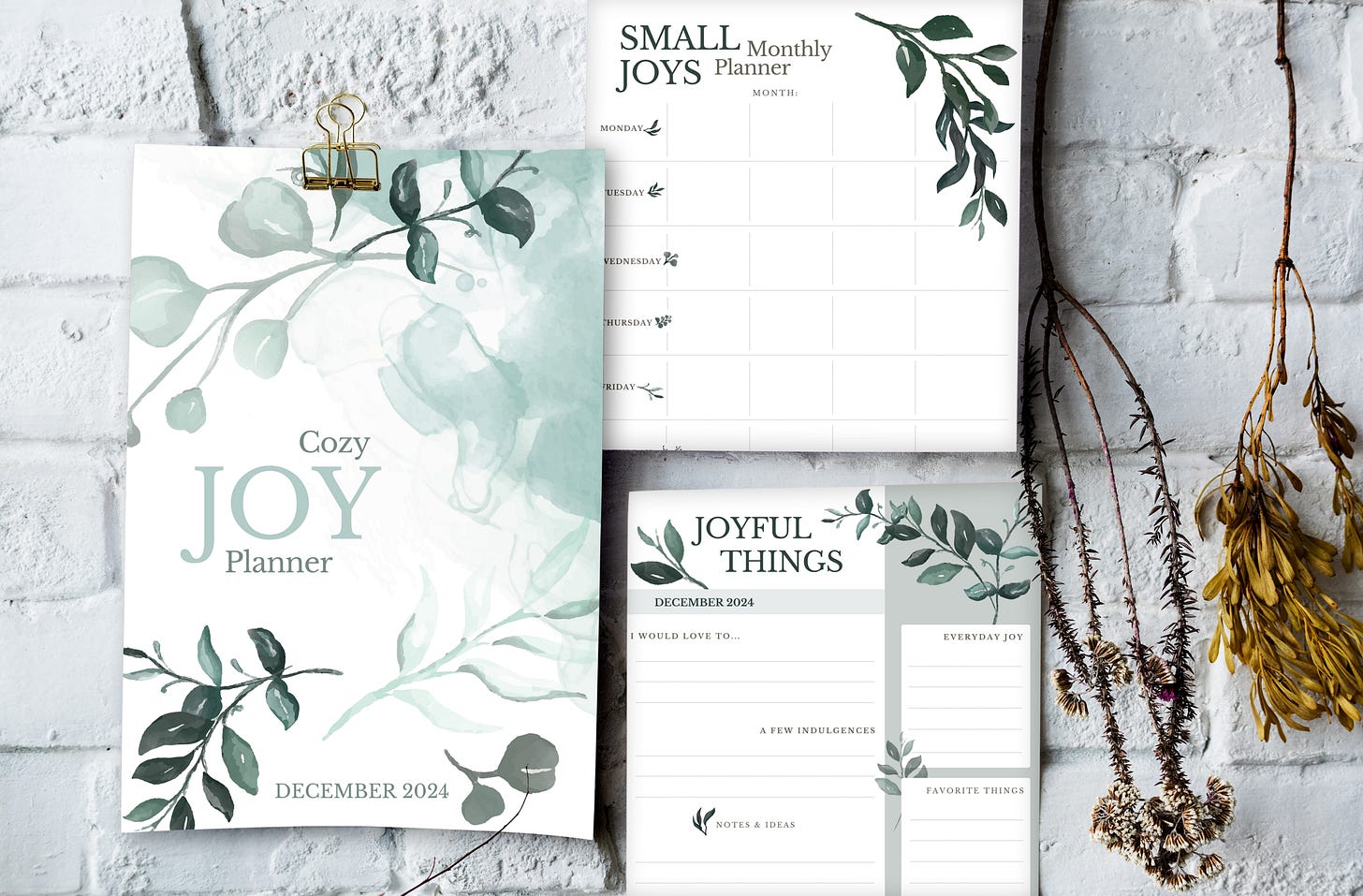 Three pages of the Joy Planner hanging on a white brick wall, surrounded by pretty dry twigs.