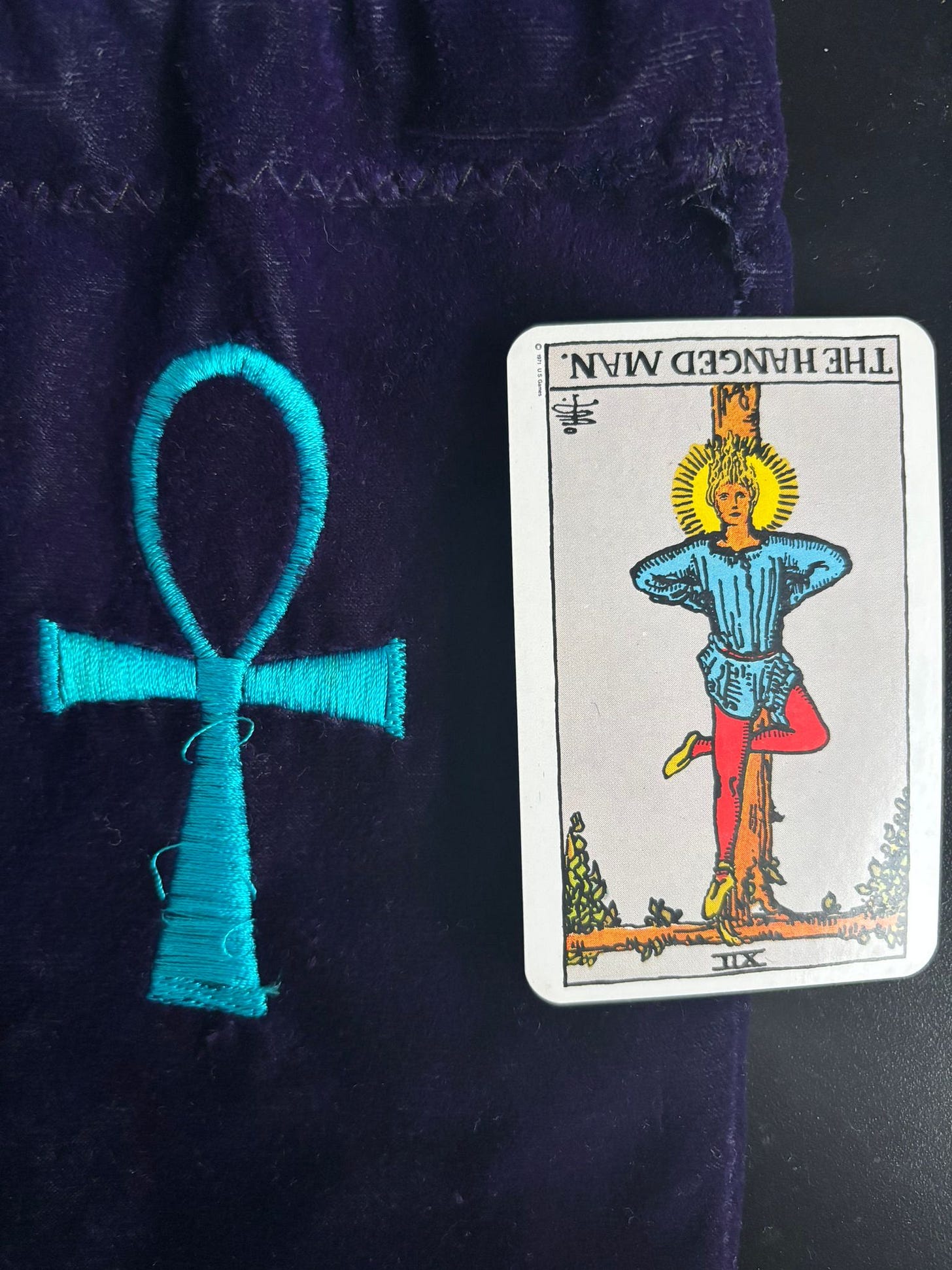 Rider-Waite tarot card “The Hanged Man” shown upside down beside a turquoise embroidered ankh on a dark purple velvet pouch.