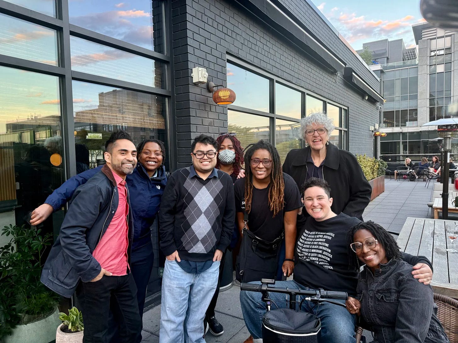 Disability Culture Lab 2025 Fellows and DCL staff pose for a group photo on an outdoor patio. Eight people smile at the camera, with one person using a power wheelchair. Modern buildings and outdoor seating are visible in the background.