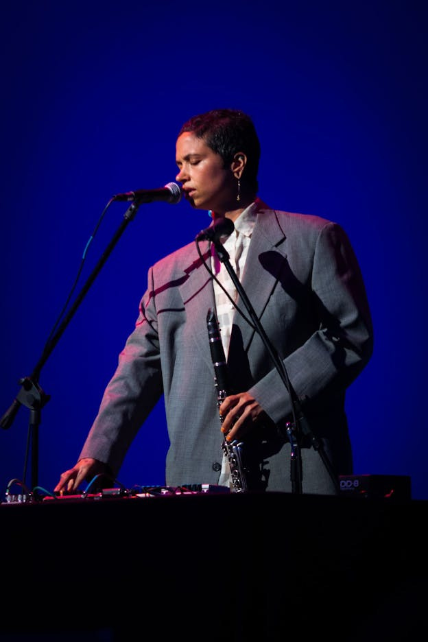Holland Andrews stands against a blue background. One hand holds a clarinet and the other rests on a mixing board. In front of them are two microphones. Their eyes are closed and their head is faced downward. They are wearing a button down top with an unbuttoned grey textured blazer. Holland Andrews stands against a blue background. One hand holds a clarinet and the other rests on a mixing board. In front of them are two microphones. Their eyes are closed and their head is faced downward. They are wearing a button down top with an unbuttoned grey textured blazer.