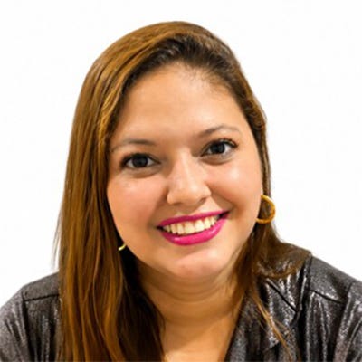 Uma pessoa está posicionada de frente para a câmera, inclinando levemente o corpo para a frente e sorrindo de forma aberta. Tem cabelos longos, lisos, castanhos, repartidos de lado.Veste uma blusa de mangas compridas em tecido brilhante, em tom escuro, com decote em "V" e detalhes pretos na região do peito. Também usa brincos de argola dourados e um colar. O fundo é liso, branco e sem elementos. Uma pessoa está posicionada de frente para a câmera, inclinando levemente o corpo para a frente e sorrindo de forma aberta. Tem cabelos longos, lisos, castanhos, repartidos de lado.Veste uma blusa de mangas compridas em tecido brilhante, em tom escuro, com decote em "V" e detalhes pretos na região do peito. Também usa brincos de argola dourados e um colar. O fundo é liso, branco e sem elementos.