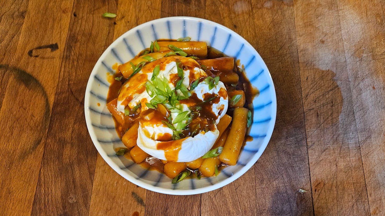 a small, shallow, white bowl with blue stripes is full of spicy tteokbokki topped with torn burrata cheese and green onions on a wooden cutting board