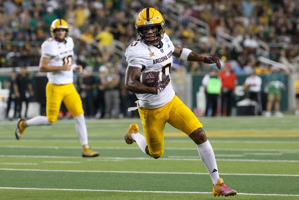 Jordan Tyson, wearing a white Arizona State jersey, yellow pants, and a yellow helmet, with high white socks holds the football in his right arm and sprints fast up the field.