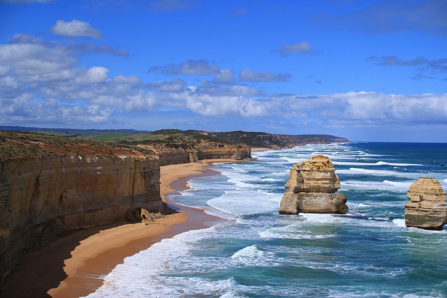Rocky outcrops can be seen off a long coast flanked by tall orange cliffs against a blue sky