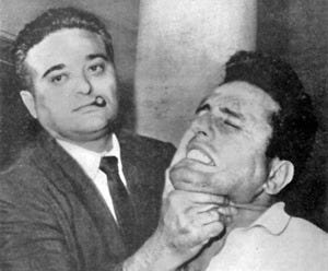 A black-and-white photograph showing a man wearing a jacket and tie with a short sigar in his mouth, on the left, holding up the chin of a man on the right, who appears to be in pain A black-and-white photograph showing a man wearing a jacket and tie with a short sigar in his mouth, on the left, holding up the chin of a man on the right, who appears to be in pain
