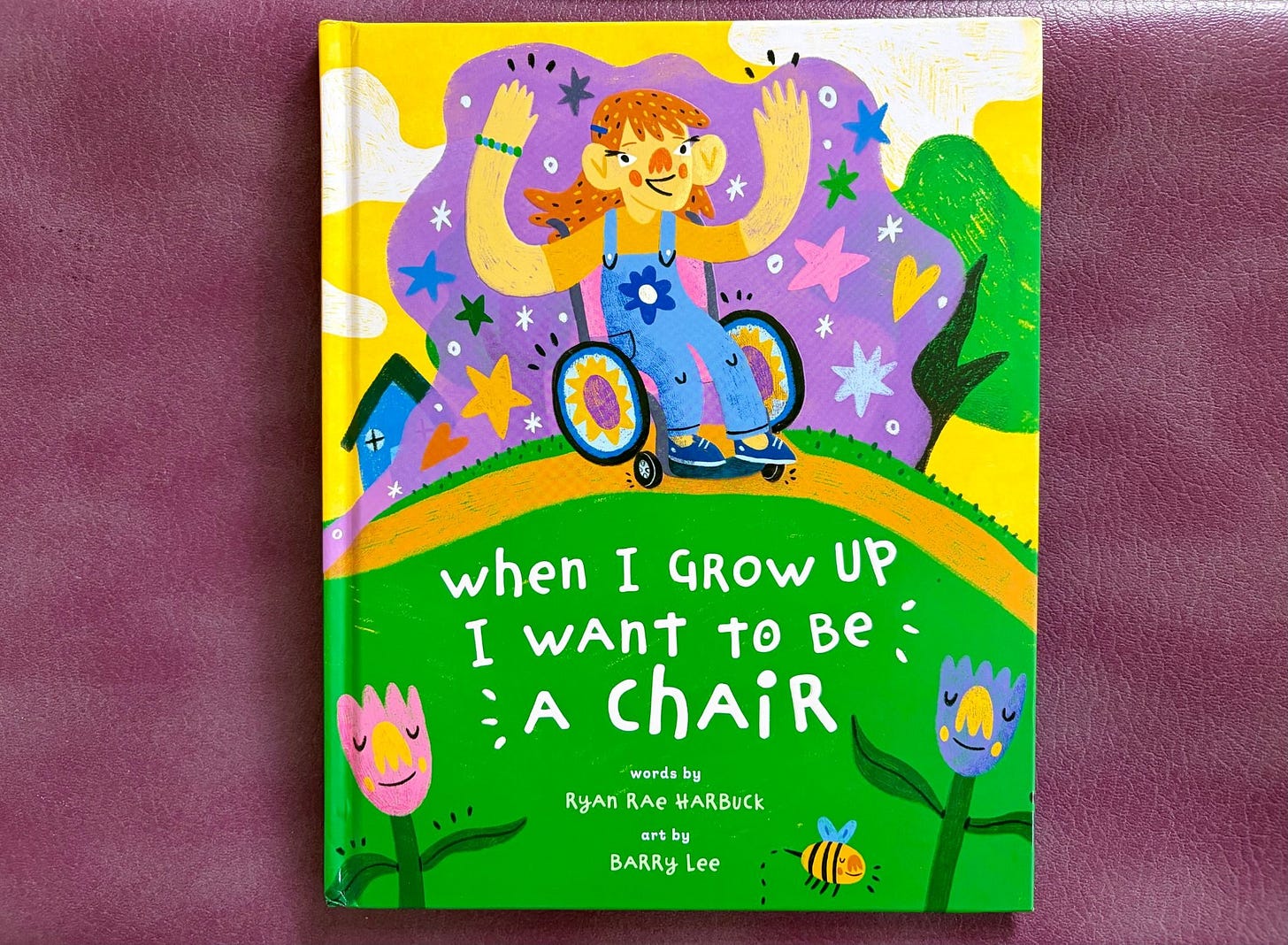 The cover of a bright and vibrant picture book against a purple background. The top features a kid sitting in a wheelchair with her arms up. Her wheels are decorated with flower covers. Behind her are stars and hearts and sparkles in front of a purple background along with a house and tree. She's sitting on top of a green hill, which feature the white words against green "When I Grow Up I Want to Be a Chair." There are two flowers with little yellow noses in each bottom corner and a cute little bee.