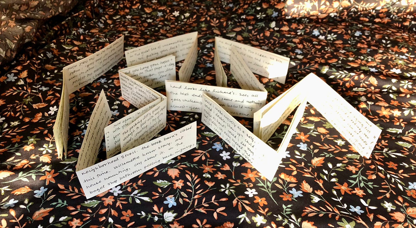 a collection of paper scraps, folded and written on in black ink, arranged to create a maze-looking pattern atop dark floral bedsheets