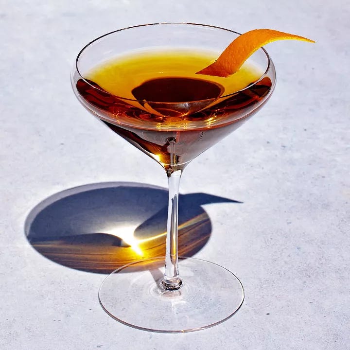 A cocktail class with a rounded, tapered lip holds a vividly gold drink garnished with a slice of orange peel. The glass rests on a brightly lit gray surface. A cocktail class with a rounded, tapered lip holds a vividly gold drink garnished with a slice of orange peel. The glass rests on a brightly lit gray surface.