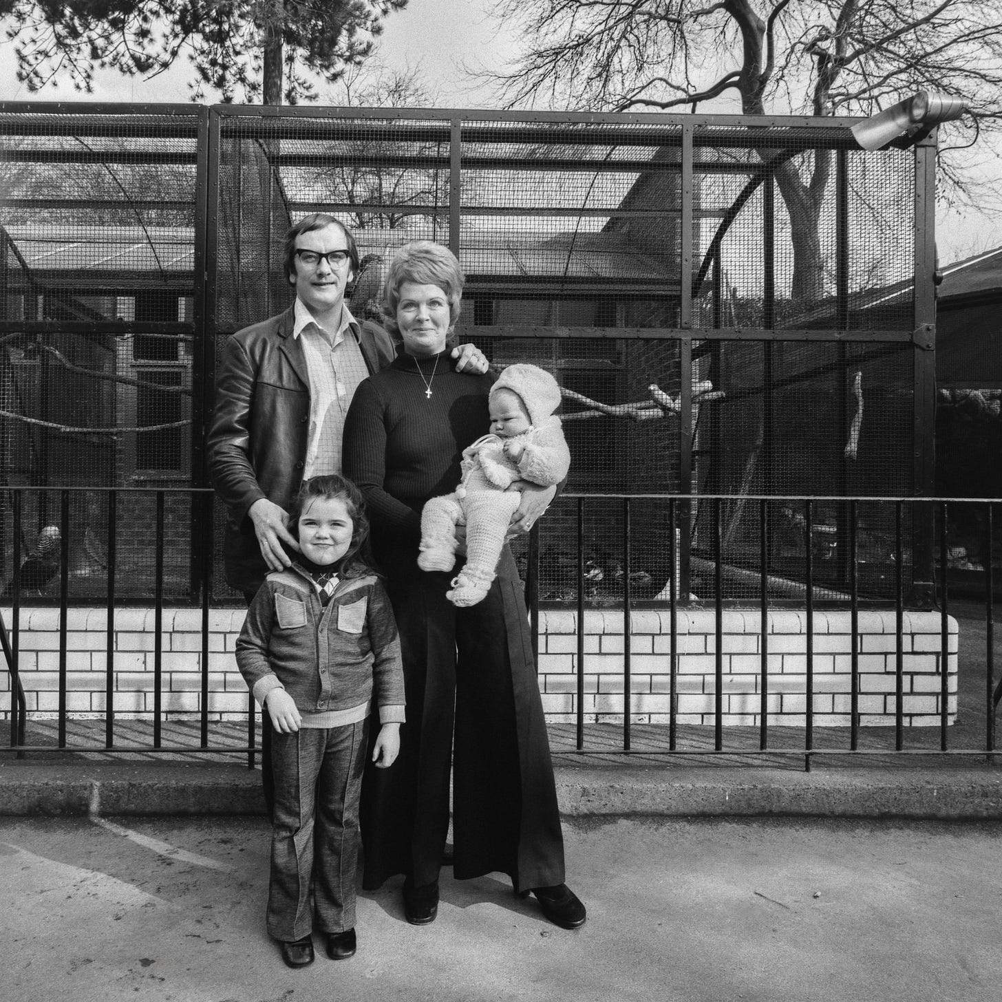 A family of four - a man, woman, child, and baby - standing in front of outdoor bird cages, with trees behind them A family of four - a man, woman, child, and baby - standing in front of outdoor bird cages, with trees behind them