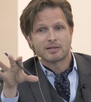 First image shows a man with light brown hair wearing a light blue shirt, dark gray suit jacket, and patterned tie, speaking into a microphone with a yellow earpiece and holding a pen in his right hand against a plain light background. Second image depicts several men in a nighttime outdoor setting with green and white lights, one wearing a green headband holding a large green flag, others in dark clothing holding and waving a large American flag over an intense fire on the ground.