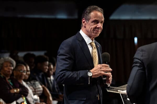 Mr. Cuomo, seated, prepares to answer a question, his hand grazing the top of a microphone.