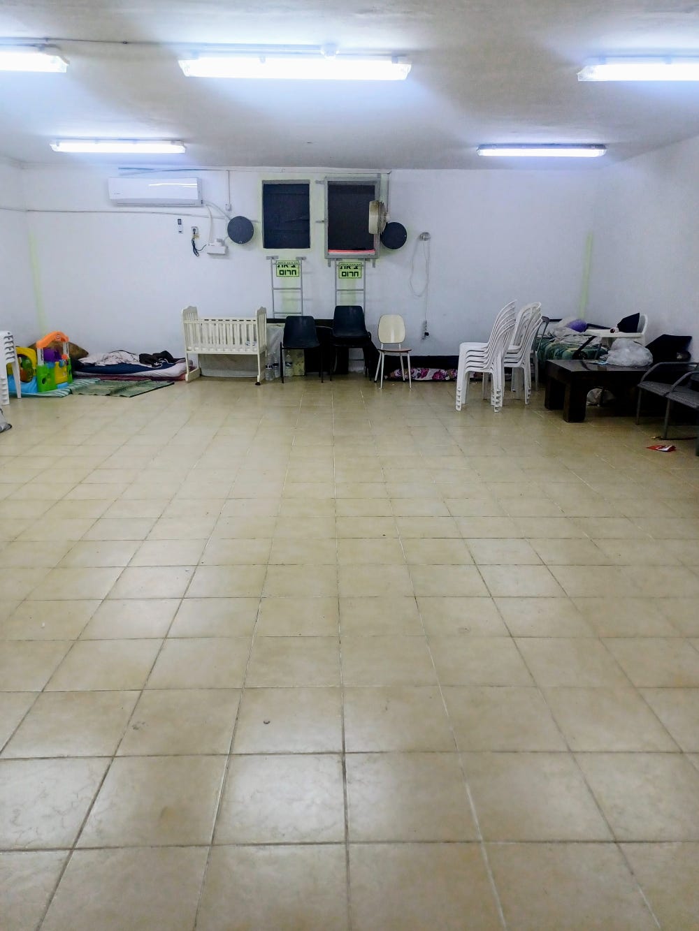 A large white-painted room with an old tile floor, walls lined with assorted chairs, a daybed, mattress with blankets, crib, and child's toys A large white-painted room with an old tile floor, walls lined with assorted chairs, a daybed, mattress with blankets, crib, and child's toys