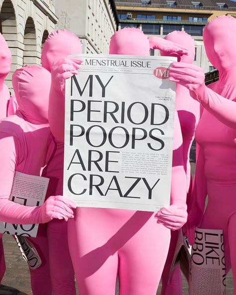 A group of people dressed in bright pink full-body suits holding newspapers titled "The Menstrual Issue," with bold headlines such as "I GET BUTT CRAMPS ON MY PERIOD" and "IT'S OFFICIAL YOU'RE NOT THE ONLY ONE," standing outdoors in an urban setting.  Individuals in bright pink full-body suits sitting and leaning against a building reading newspapers titled "The Menstrual Issue," with bold headlines like "BIG PADS ARE MY HERO."  A group of people in vibrant pink full-body suits holding a newspaper prominently displaying the bold headline "MY PERIOD POOPS ARE CRAZY," outdoors in front of city buildings.