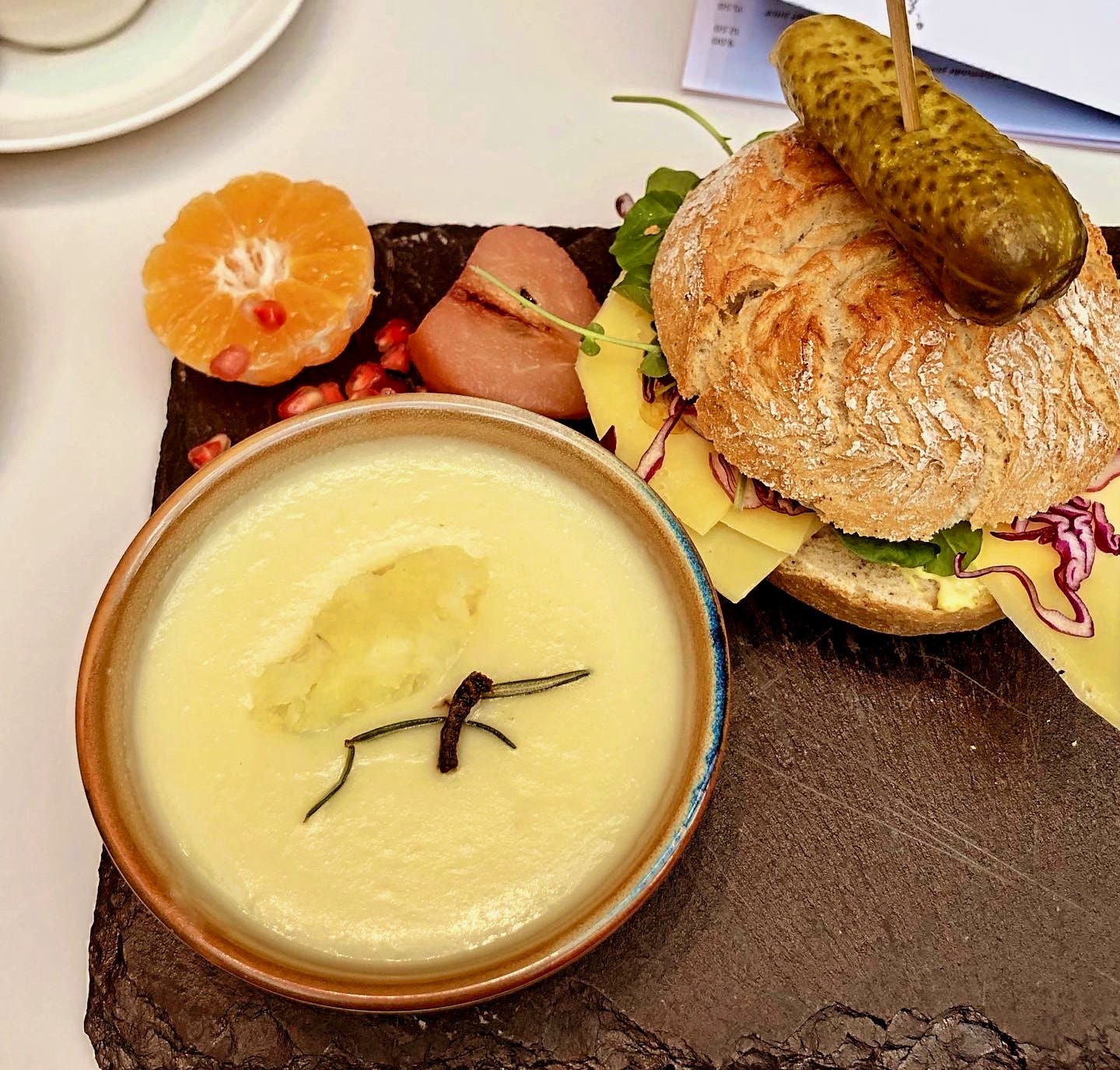 Lunch at the art museum cafe: a small bowl of parsnip soup topped with roasted herbs, a crusty white roll of cheese and red cabbage, topped with a large gherkin. Served on a black slate, which also features a half orange, half a poached pear, and a pile of pomegranate seeds. The plating is intended to evoke the atmosphere of a classic Dutch "still life" painting.