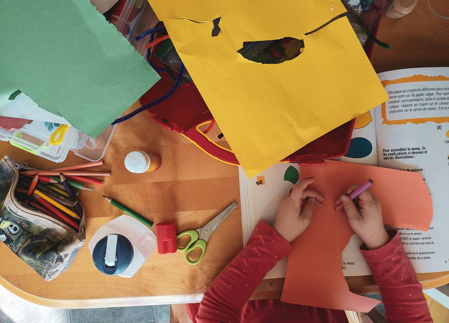 Overhead view of a child's hands working on a paper craft project at a wooden table, surrounded by colored pencils, safety scissors, a glue bottle, a pencil sharpener, and sheets of orange, yellow, and green construction paper. An open craft activity book with French instructions is visible to the right.