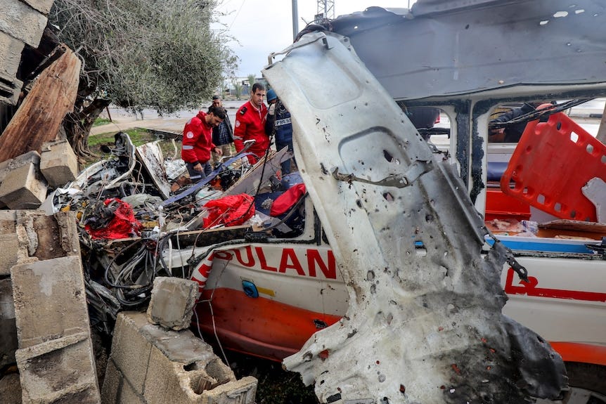 A medium shot of a group of medics in uniform standing near a destroyed ambulance van and rubble.
