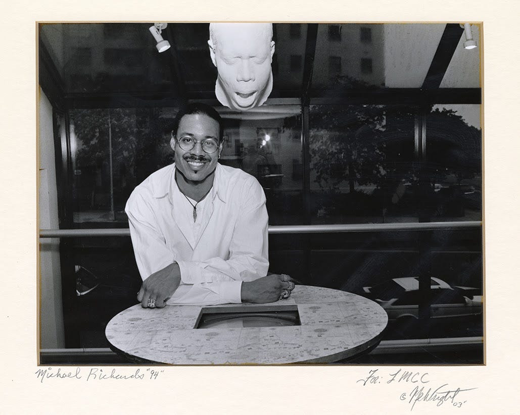 A black and white self portrait of Michael Richards, a black masc presenting person sitting with their arms crossed at a table in 1990s new york city wearing a white long sleeve shirt. A white sculpture of a face with an open mouth hangs above them.