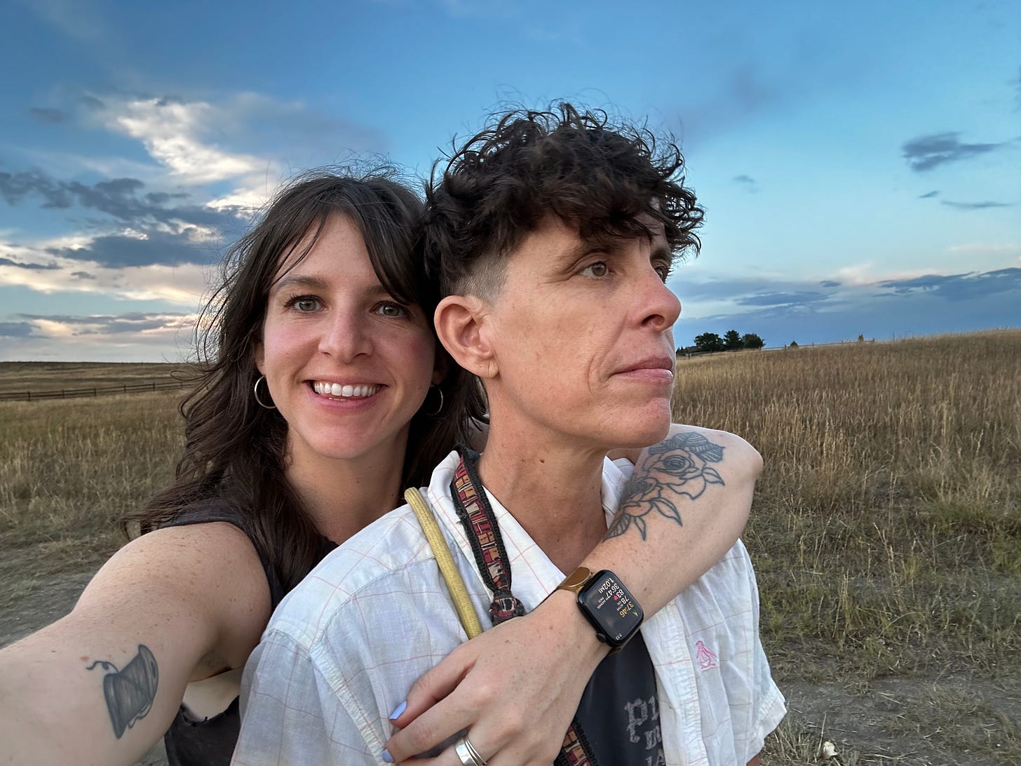 Meg smiles at the camera with her arm around Andrea, as Andrea looks knowingly off into the distance. The two lovers are backdropped by the plains and big blue skies of Colorado