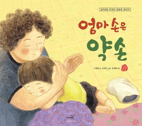 A warm and tender illustration of a Korean children's book titled "Mom's Hand is a Healing Hand". The cover shows an affectionate mother figure with curly hair holding a young child close, symbolizing the comfort and healing power of a mother's touch. The title is written in soft, colorful Korean characters.