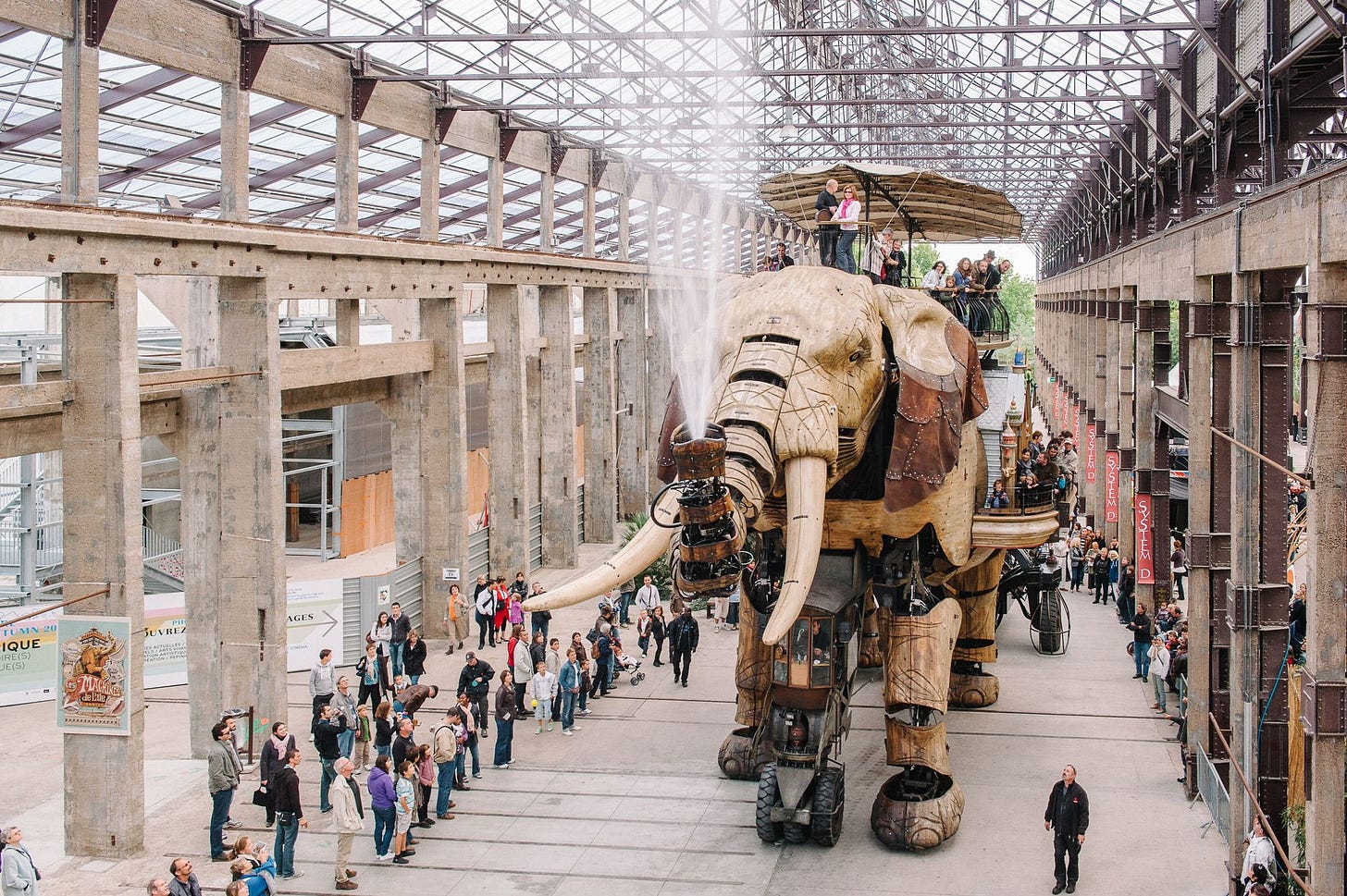 A giant wooden elephant carrying people walks around an empty warehouse while spraying water from its trunk.